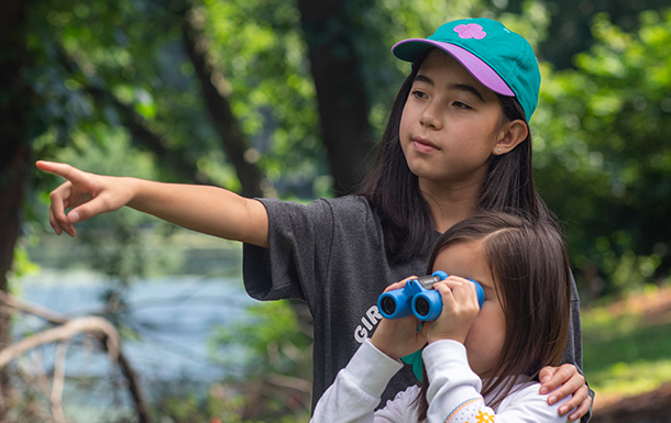 two young girl scouts outside at state park using binoculars - older girl is helping daisy girl scout see nature